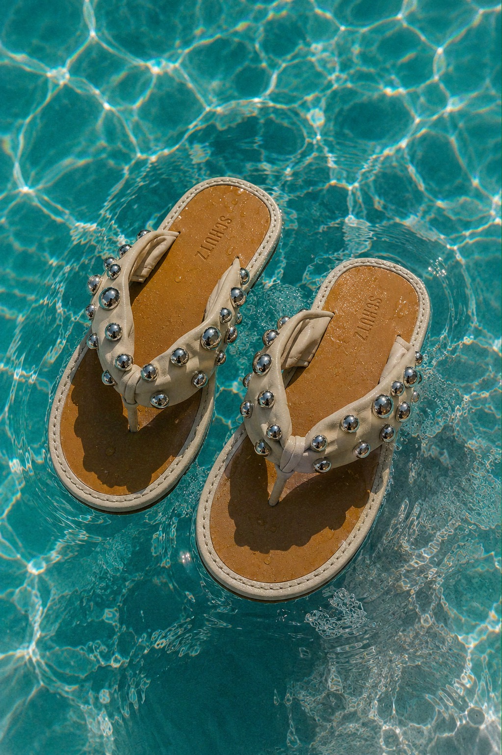 Chinelos SCHUTZ com aplicações metálicas flutuando na piscina