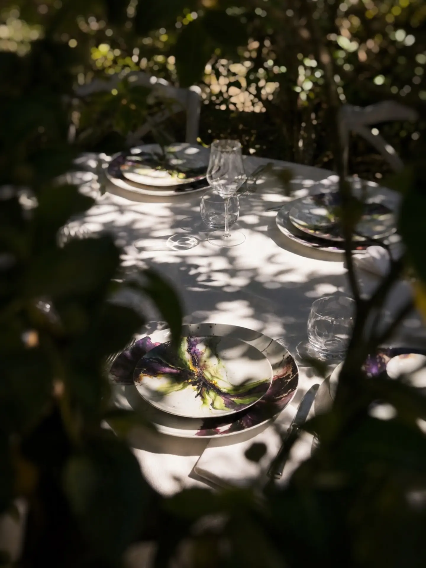 Mesa posta ao ar livre com porcelanas pintadas à mão entre luz e sombras da vegetação